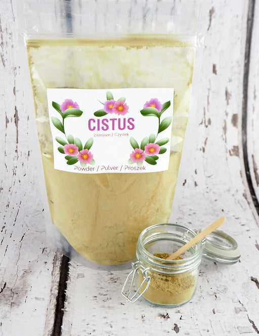 Cistus-Kräutertee in Pulverform in einer transparenten Tüte, präsentiert auf einem Holztisch neben einem offenen Glasgefäß mit Pulver und Holzlöffel.