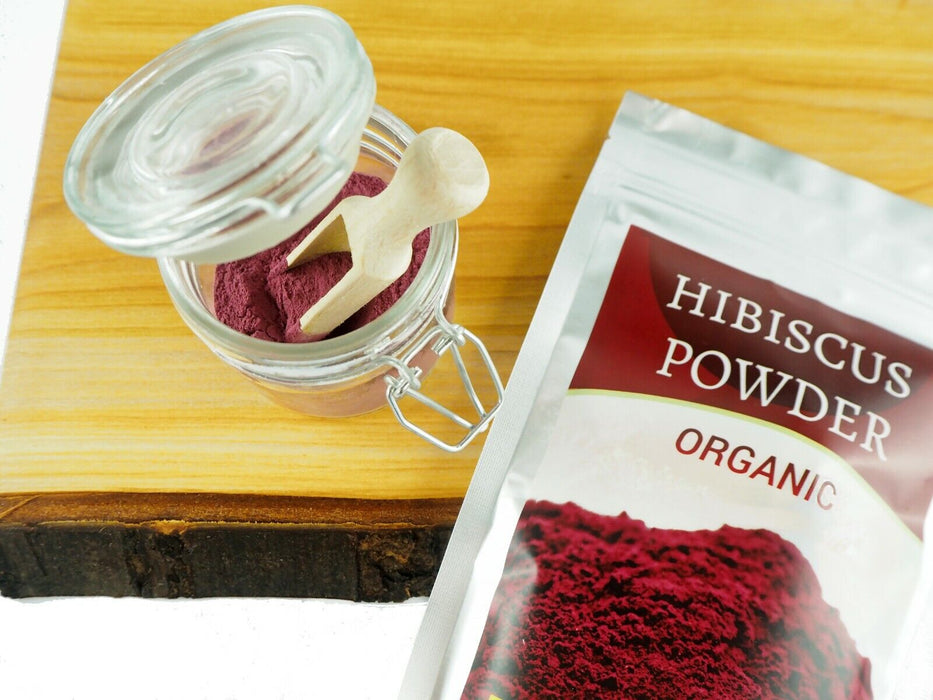 Ein Glasgefäß mit biologischem Hibiskuspulver und einem hölzernen Messlöffel, daneben eine Verpackung mit der Aufschrift 'HIBISCUS POWDER ORGANIC