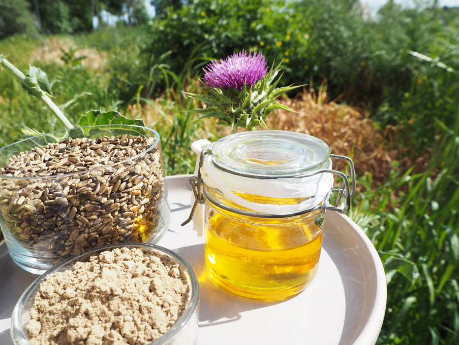 Ein Glasgefäß mit goldenem Mariendistelöl neben einer Schüssel mit Mariendistelsamen, im Hintergrund ein Feld.