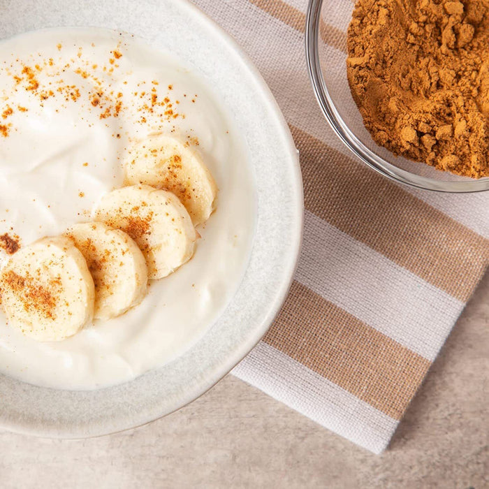 Hagebuttenpulver, sanft über cremigen Joghurt mit Bananenscheiben gestreut, neben einem Glasbehälter mit mehr Pulver und einem gestreiften Tuch im Hintergrund