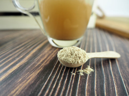 Holzlöffel mit natürlichem Pflanzenpulver vor einer Tasse mit goldbrauner Flüssigkeit auf einem dunkel gestreiften Holztisch.